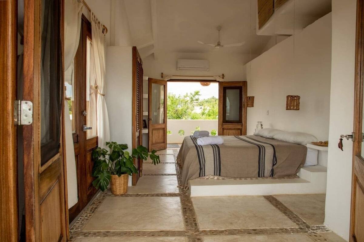 A bedroom with a bed, a potted plant, a ceiling fan, and a view of greenery outside.
