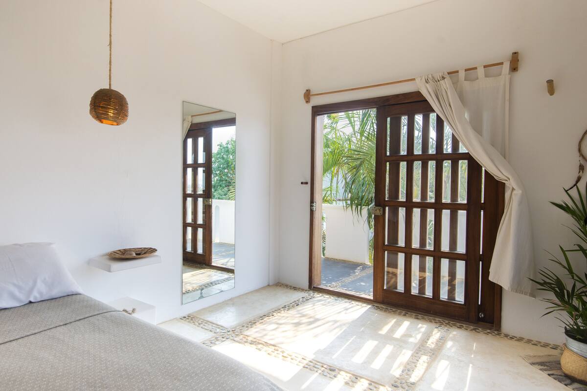 A bedroom with a bed, a hanging light fixture, a small shelf, a mirror, and a sliding glass door leading to an outdoor area.