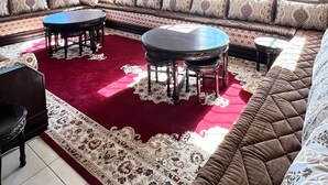 Un salon spacieux avec un grand canapé d’angle, deux tables rondes et un tapis rouge à motifs.