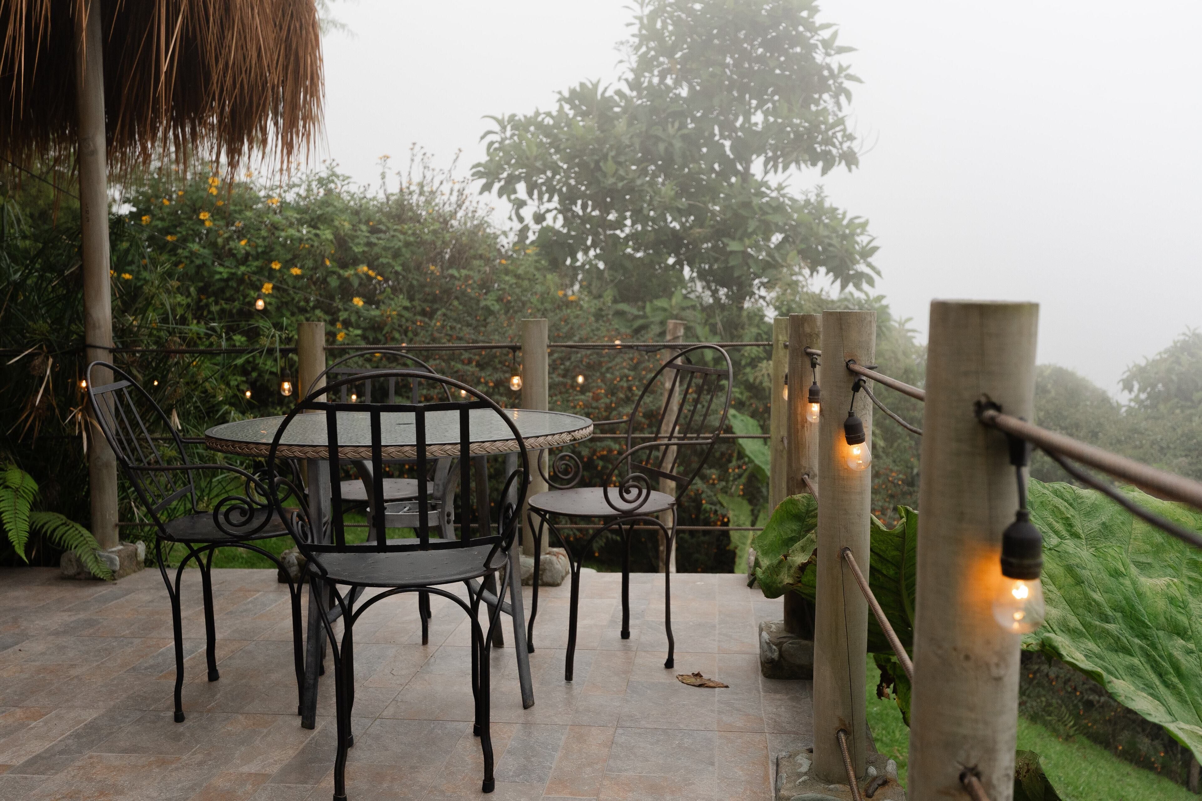 Un patio al aire libre con una mesa redonda y sillas, rodeado de follaje y luces colgantes.