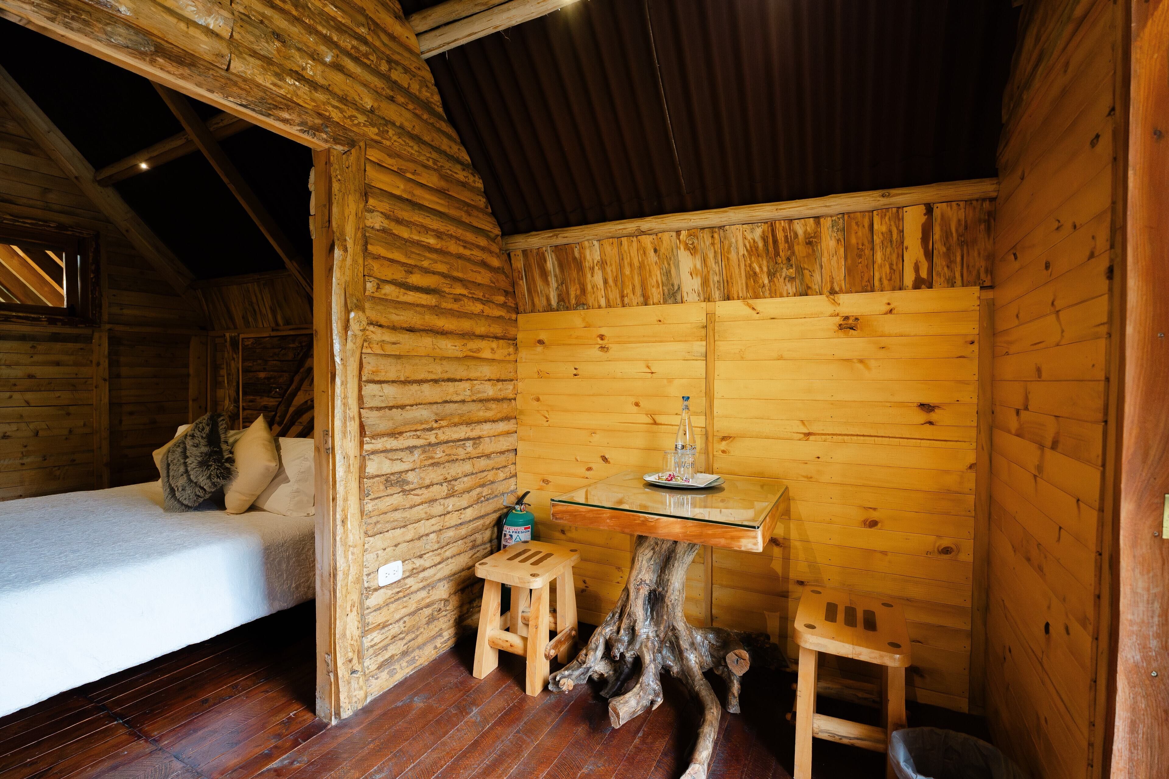 Une pièce de cabane en bois comprenant un lit, un petit tabouret, une table et un socle en forme de tronc d’arbre en bois.