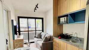 A modern kitchen with wooden cabinets, a built-in microwave, and a sink. There is a dining area with a round table and chairs, and a sofa in the background. - Brera Moema NR (São Paulo)