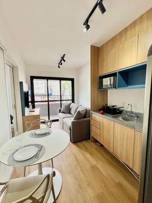A modern kitchen with wooden cabinets, a built-in microwave, and a sink. There is a dining area with a round table and chairs, and a sofa in the background. - Brera Moema NR (São Paulo)