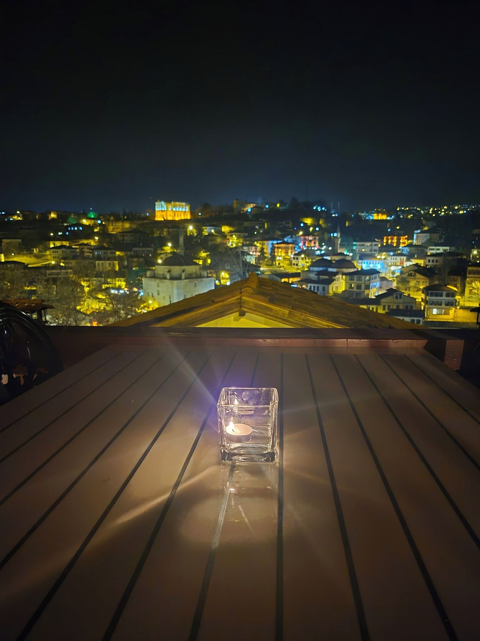 A night view from a rooftop terrace with a lit candle in the foreground, overlooking a cityscape with illuminated buildings.