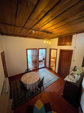 A room with a wooden ceiling, a round table with chairs, a rug, and a small table with a vase and a kettle.