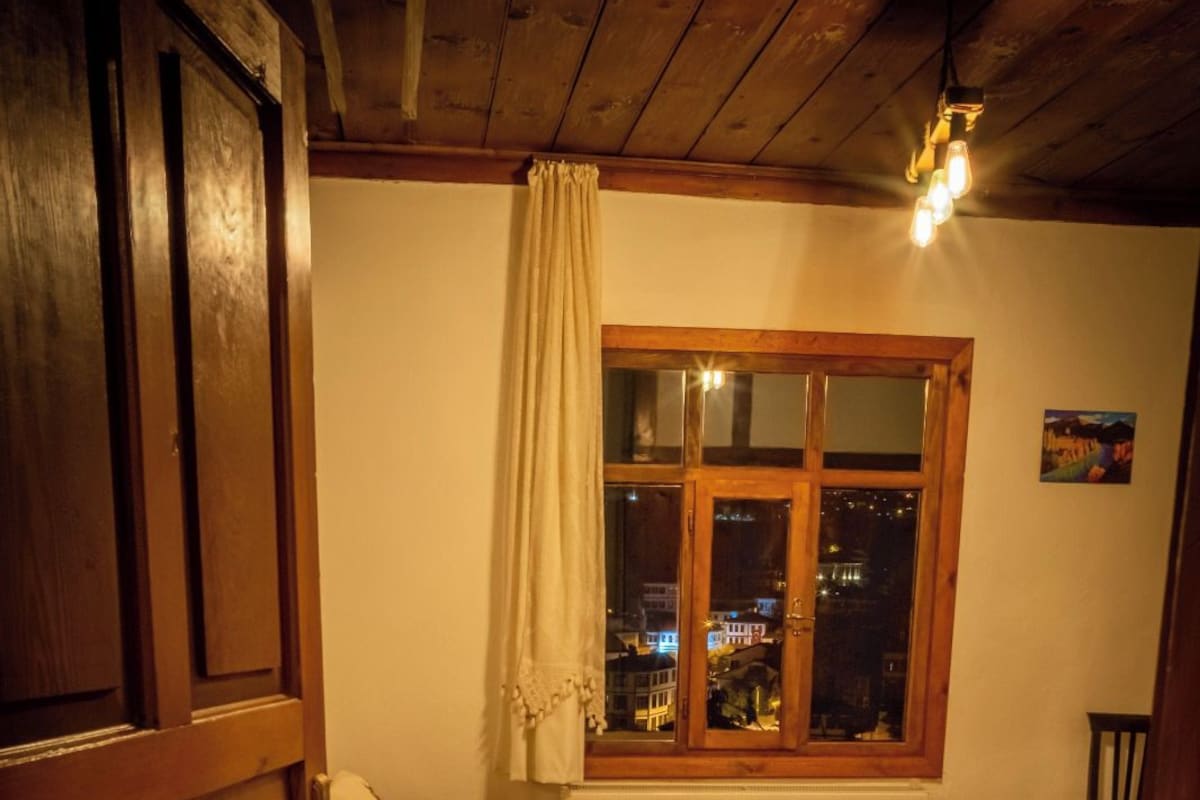 A bedroom with a wooden bed, a window with a view of the city at night, a patterned rug, and a wooden wardrobe.