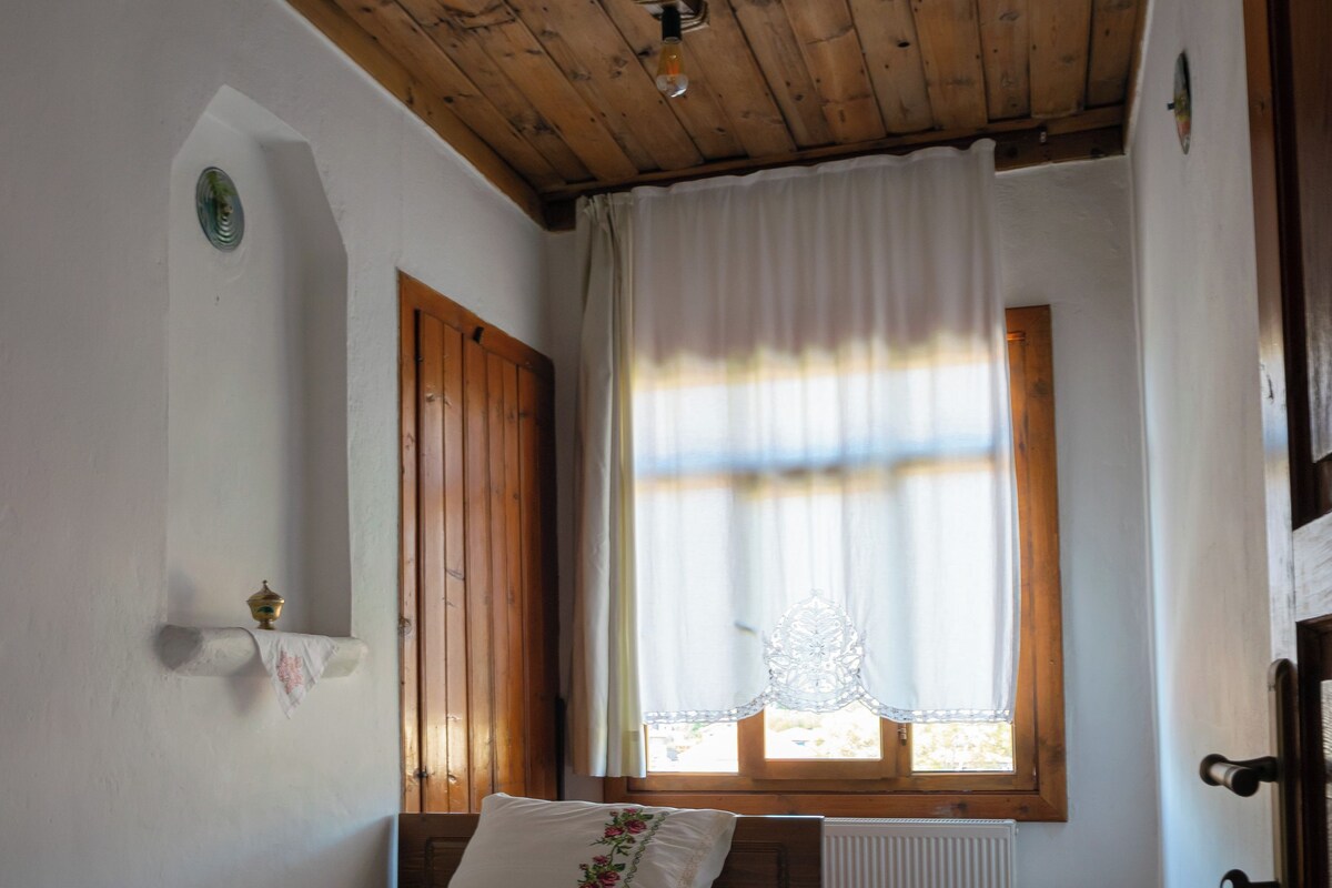 Un dormitorio con techo de madera, una cama con colcha blanca y morada, una mesita y una ventana con cortinas.