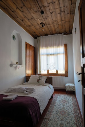 A bedroom with a wooden ceiling, a bed with a white and purple bedspread, a small table, and a window with curtains. - Meltem Hanım Konagı (Karabuk)