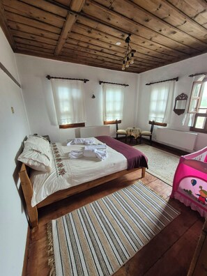 A bedroom with a wooden bed, a small table, and a chair. There are windows with curtains, a radiator, and a ceiling with exposed beams.