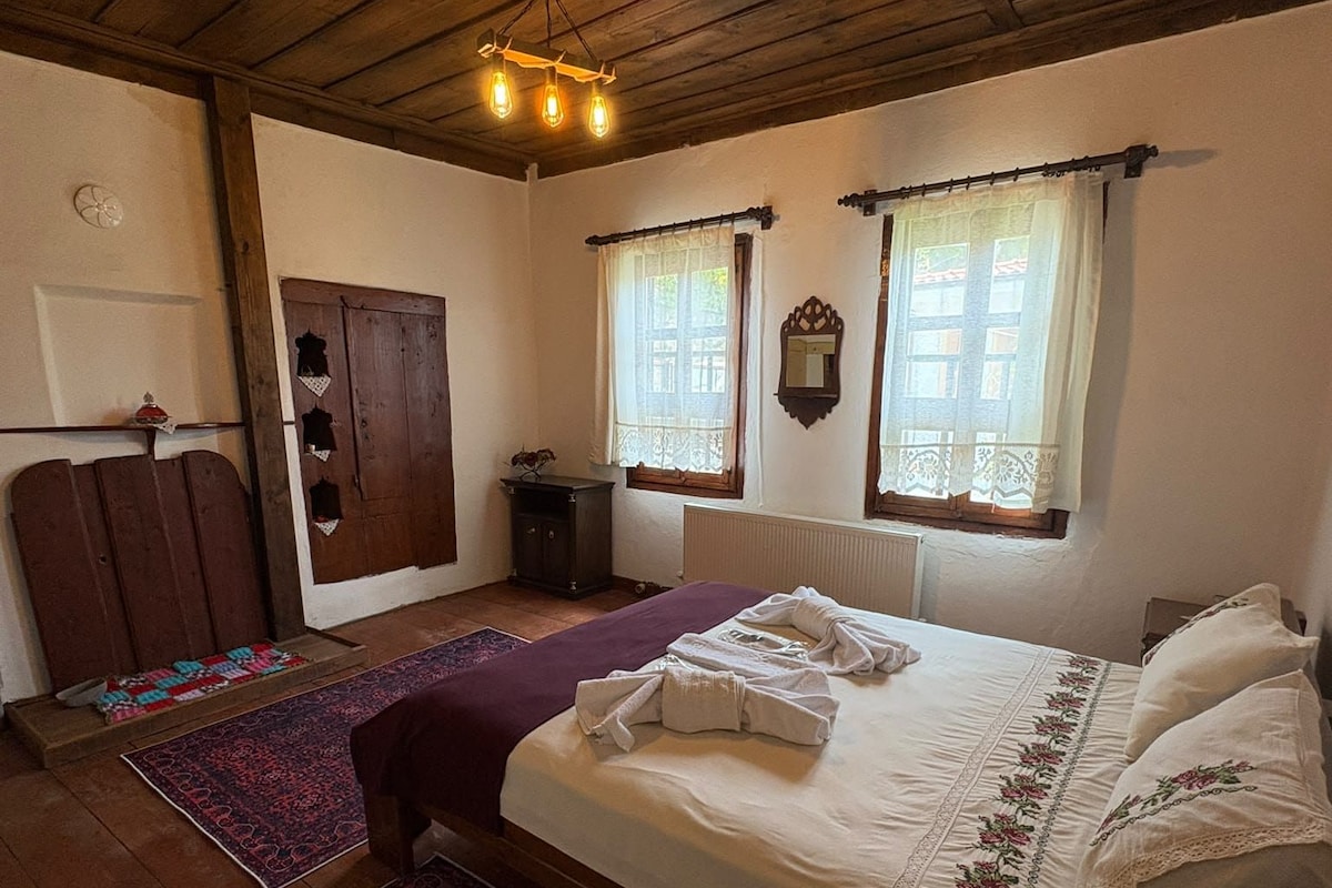 Un dormitorio con techo de madera, una cama con ropa de cama blanca, dos ventanas con cortinas, un mueble pequeño y una alfombra.