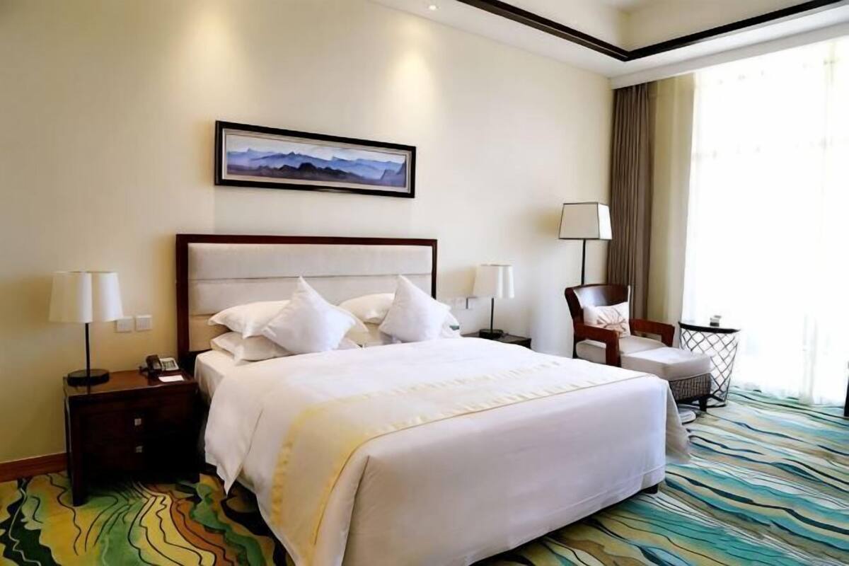 A hotel room with a large bed, bedside lamps, a chair, a desk, and a window with curtains.