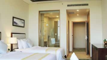 A hotel room with two beds, a desk, a lamp, a framed picture on the wall, and a bathroom visible through a glass door.