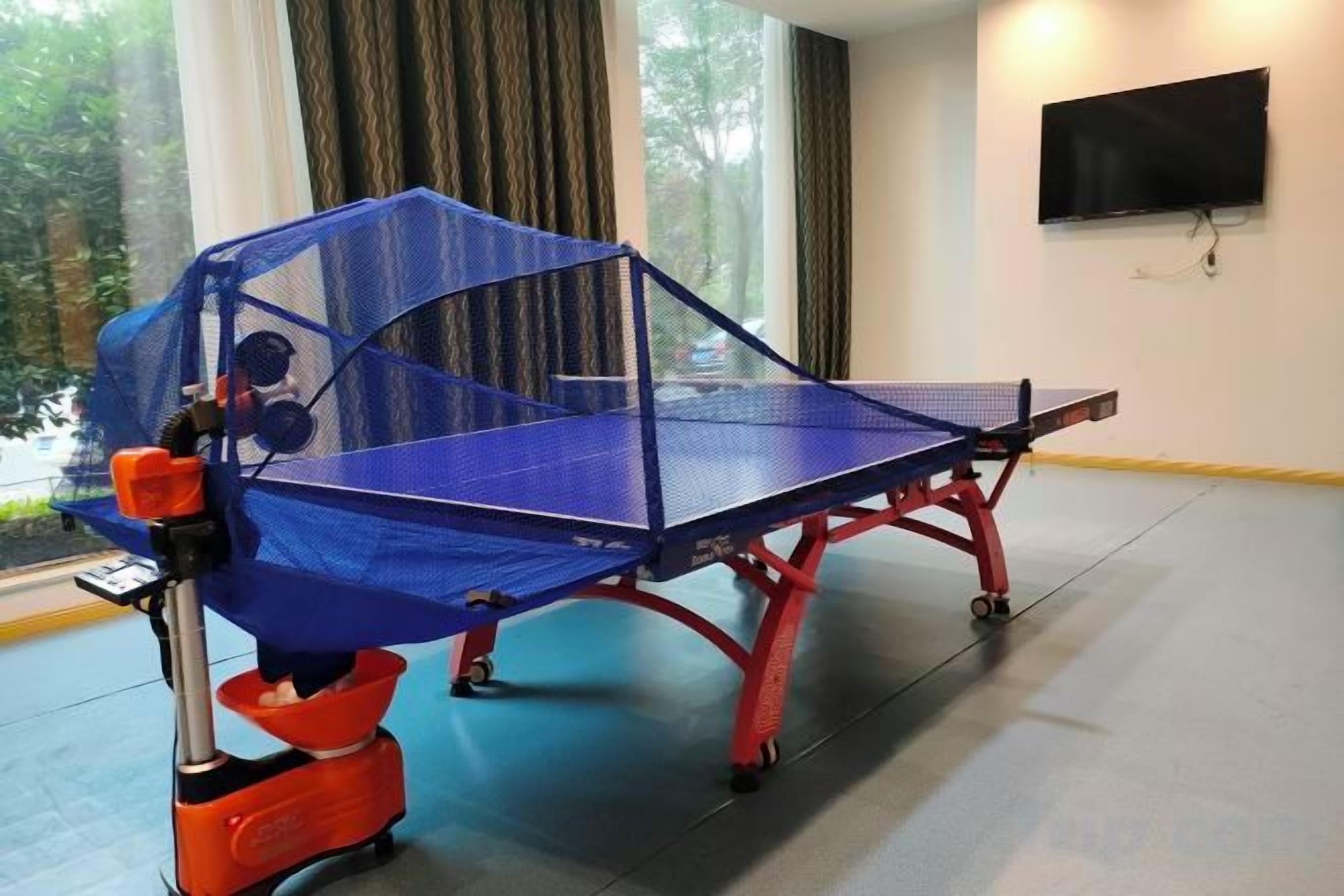 A ping pong table with a protective net and a table tennis paddle in a room with a large window and a flat-screen TV on the wall.
