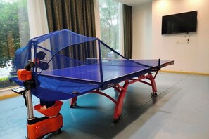 A ping pong table with a protective net and a table tennis paddle in a room with a large window and a flat-screen TV on the wall.