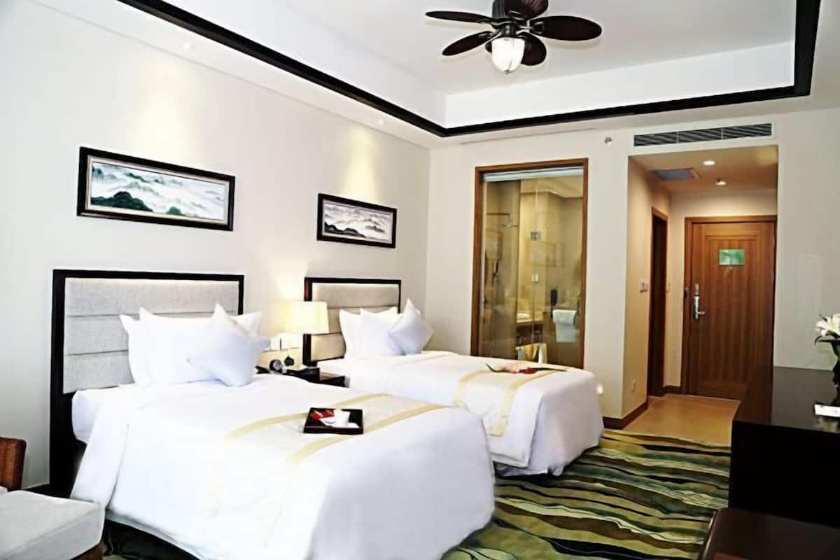 A hotel room with two beds, a ceiling fan, framed artwork on the walls, a wooden door, and a green patterned rug.