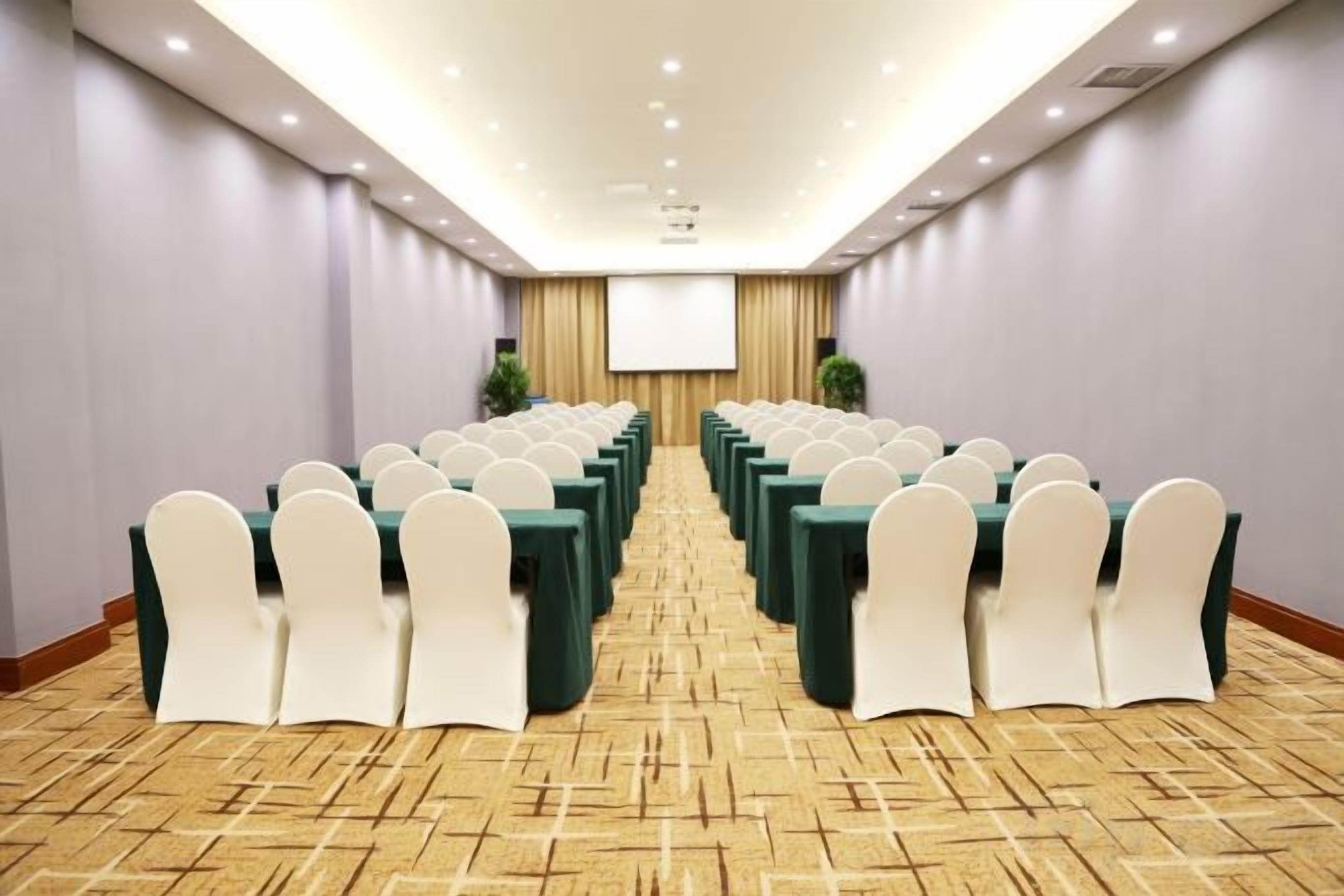 A conference room with rows of tables and chairs, a projector screen, and a carpeted floor.