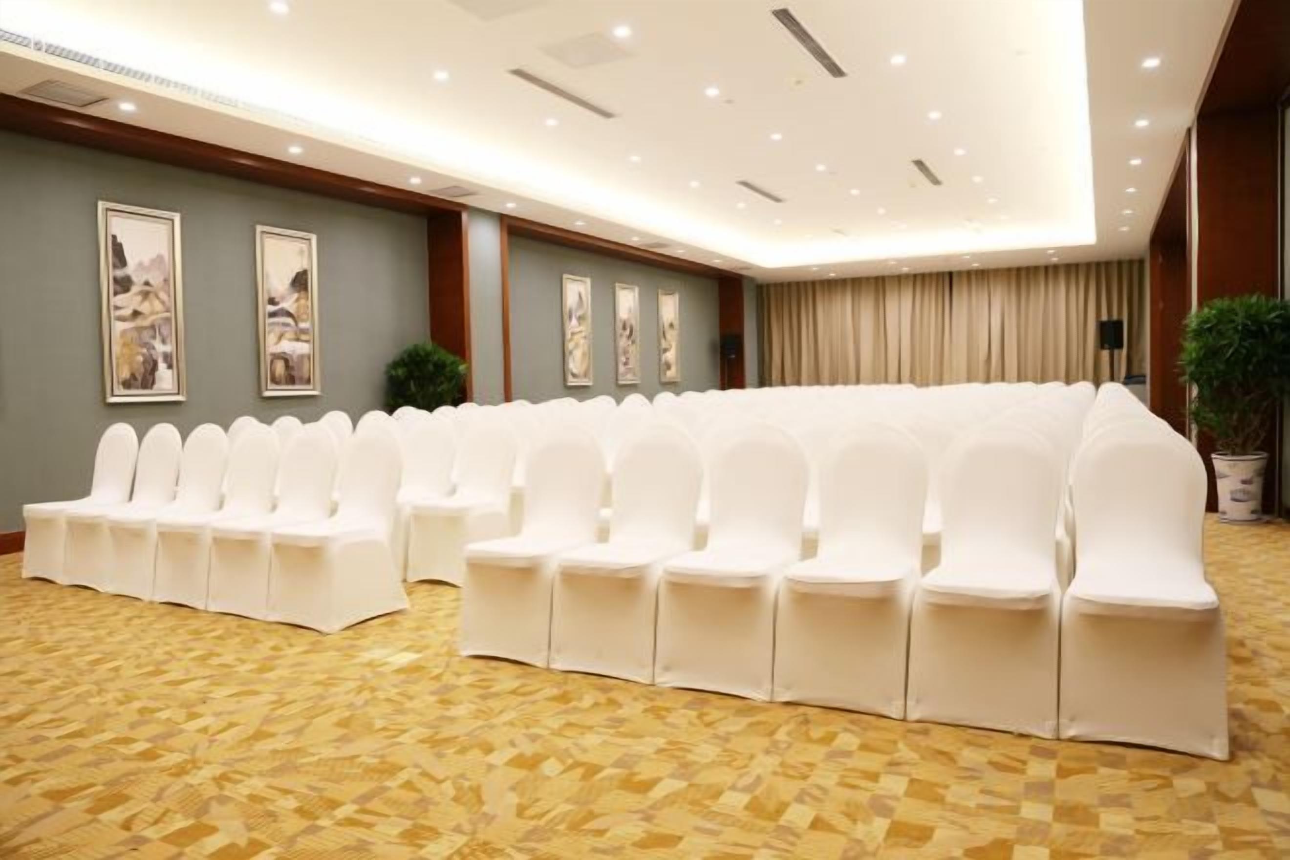 Un salón de conferencias con filas de sillas blancas, una alfombra estampada y cuadros en las paredes.