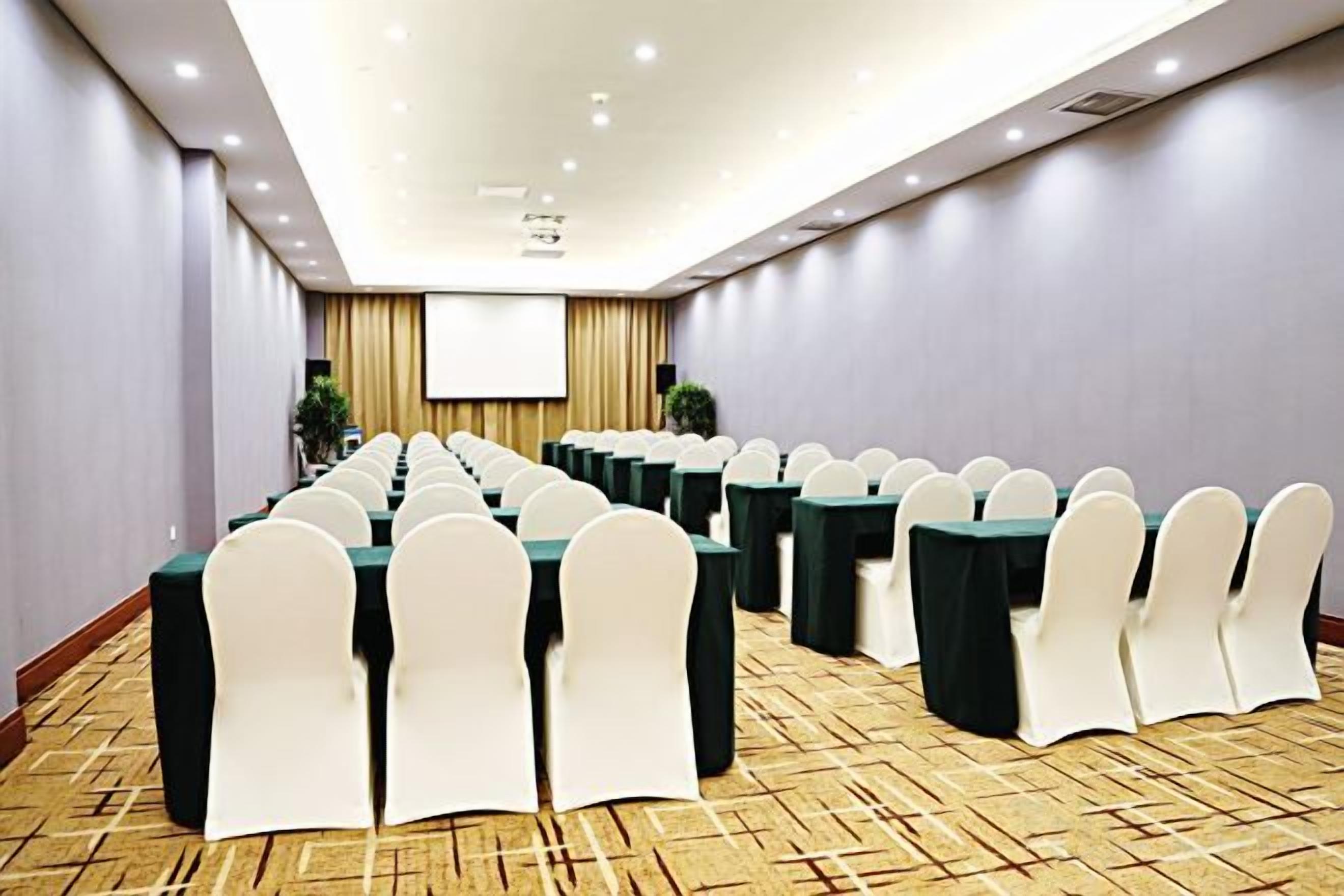 A conference room with rows of tables and chairs, a projector screen, and a carpeted floor.