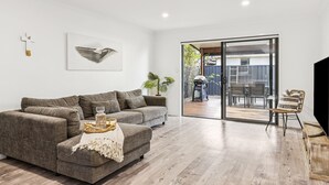A modern living room with a grey sofa, a glass coffee table, and a large sliding glass door leading to an outdoor patio area. - Beachside 24 - Pet Friendly Holiday Home - Holiday Yamba (Yamba)