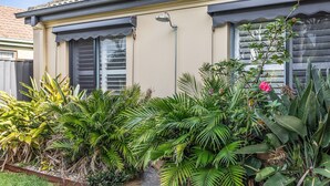 A house with a beige exterior, dark shutters, and a small garden with tropical plants. - Beachside 24 - Pet Friendly Holiday Home - Holiday Yamba (Yamba)