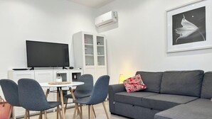 Una sala moderna con un sofá gris, una mesa de comedor con cuatro sillas, un televisor de pantalla plana y un mueble blanco con puertas de vidrio.