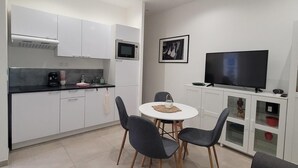 Una cocina moderna con gabinetes blancos, un microondas y una cafetera. Hay un comedor con una mesa redonda y cuatro sillas. Un televisor de pantalla plana está montado sobre un mueble blanco.