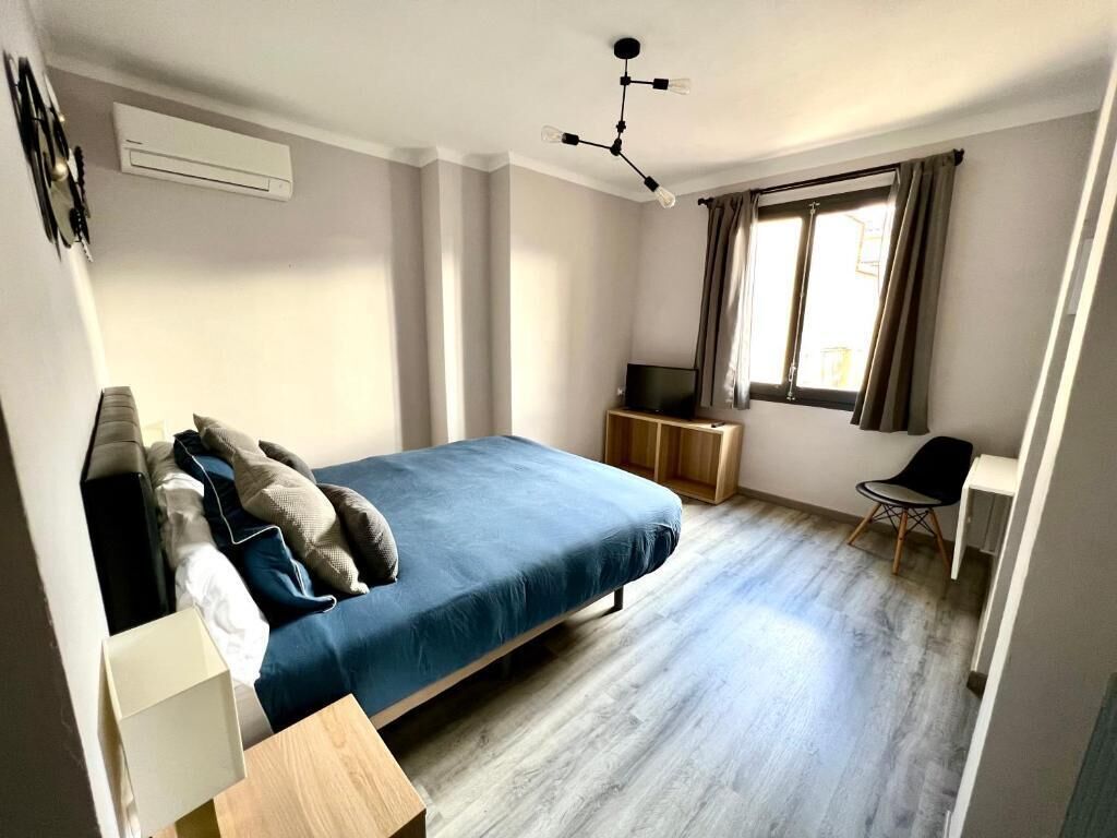 Un dormitorio con cama, escritorio, silla, aire acondicionado y ventana con cortinas.