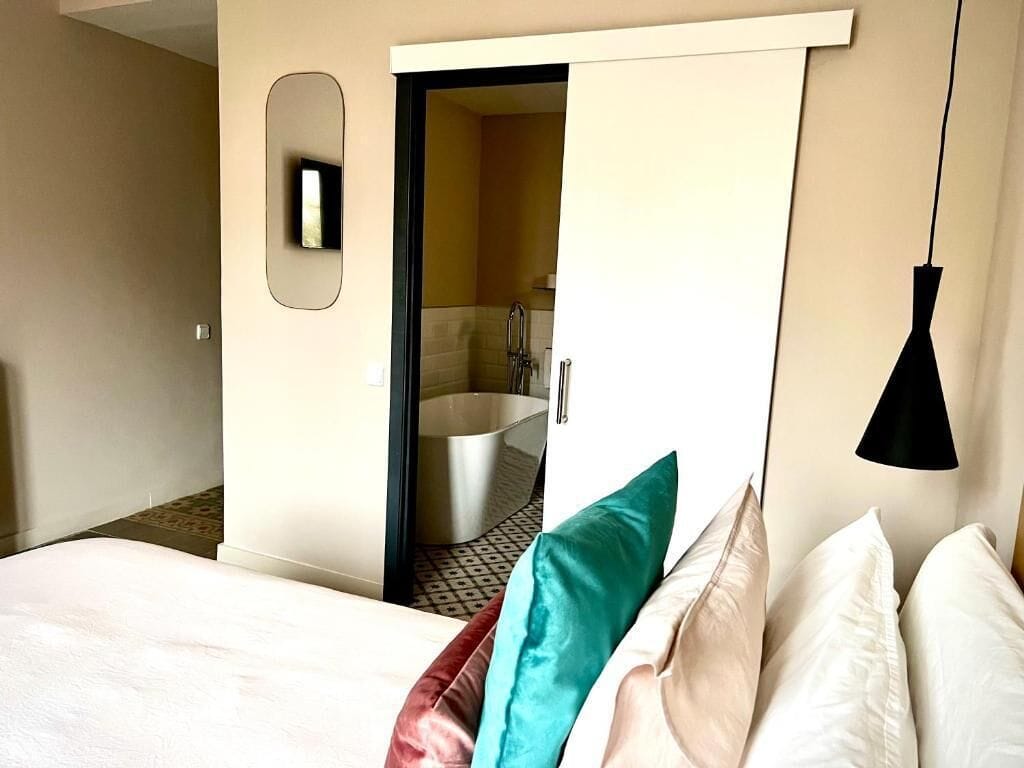 Ein Hotelzimmer mit einem Bett, einem Spiegel, einer Pendelleuchte und einer offenen Tür zu einem Badezimmer mit einer Badewanne.