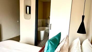 Ein Hotelzimmer mit einem Bett, einem Spiegel, einer Pendelleuchte und einer offenen Tür zu einem Badezimmer mit einer Badewanne.