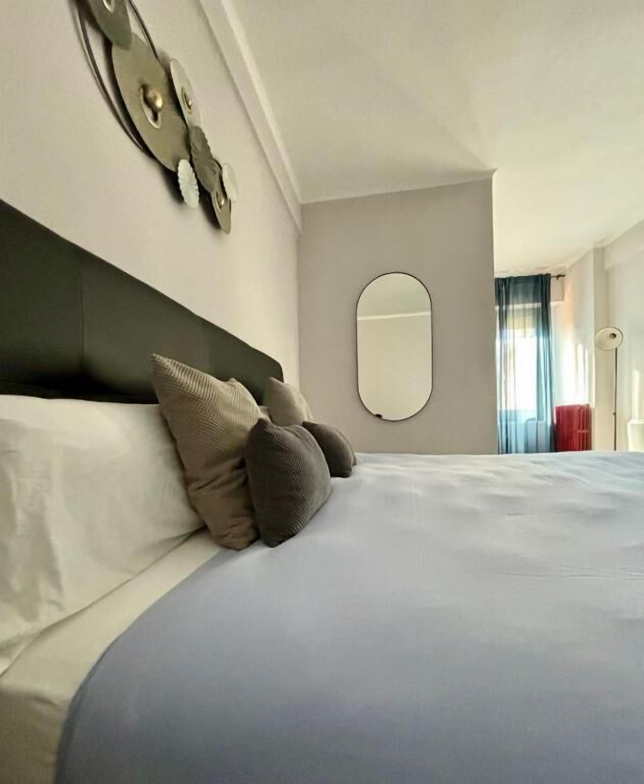 Een netjes opgemaakt bed met een blauwe deken en twee kussens, een ronde spiegel aan de muur en een raam met gordijnen.