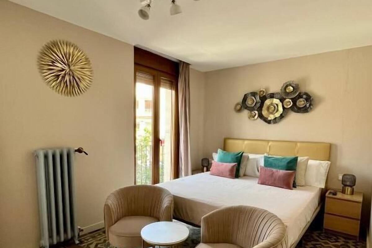 Une chambre d’hôtel avec un grand lit, deux fauteuils, une petite table, un radiateur, un élément décoratif mural et un revêtement de sol à motifs.