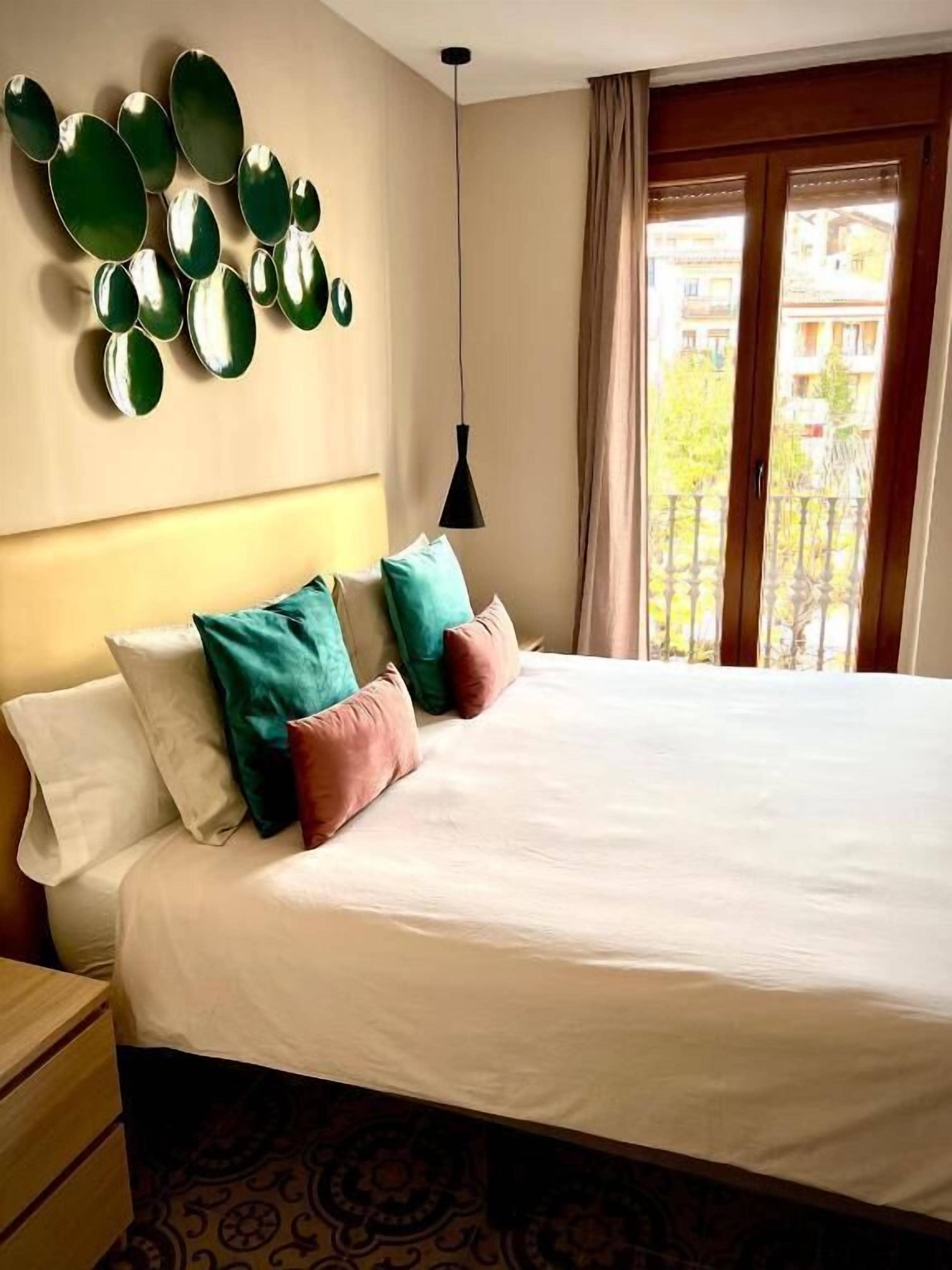 A hotel room with a bed, a nightstand, a wall-mounted mirror, a hanging pendant light, and a window with curtains.