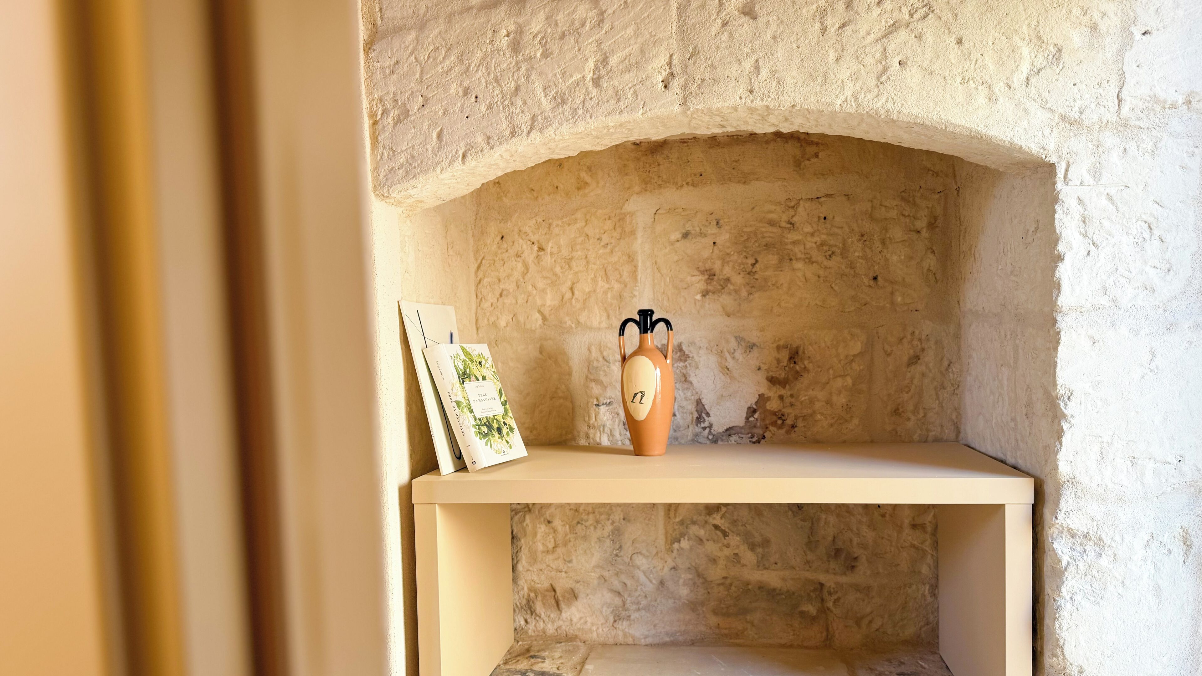 Un mur en pierre sur lequel est fixée une étagère, et sur laquelle sont posés un vase et des tableaux encadrés.