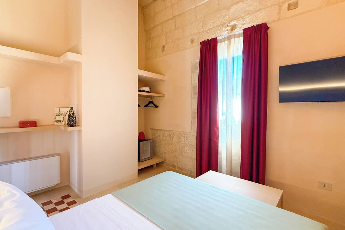 Une chambre d’hôtel avec un lit, un téléviseur fixé au mur, une petite étagère avec une boîte rouge et un vase, et une fenêtre avec des rideaux rouges.