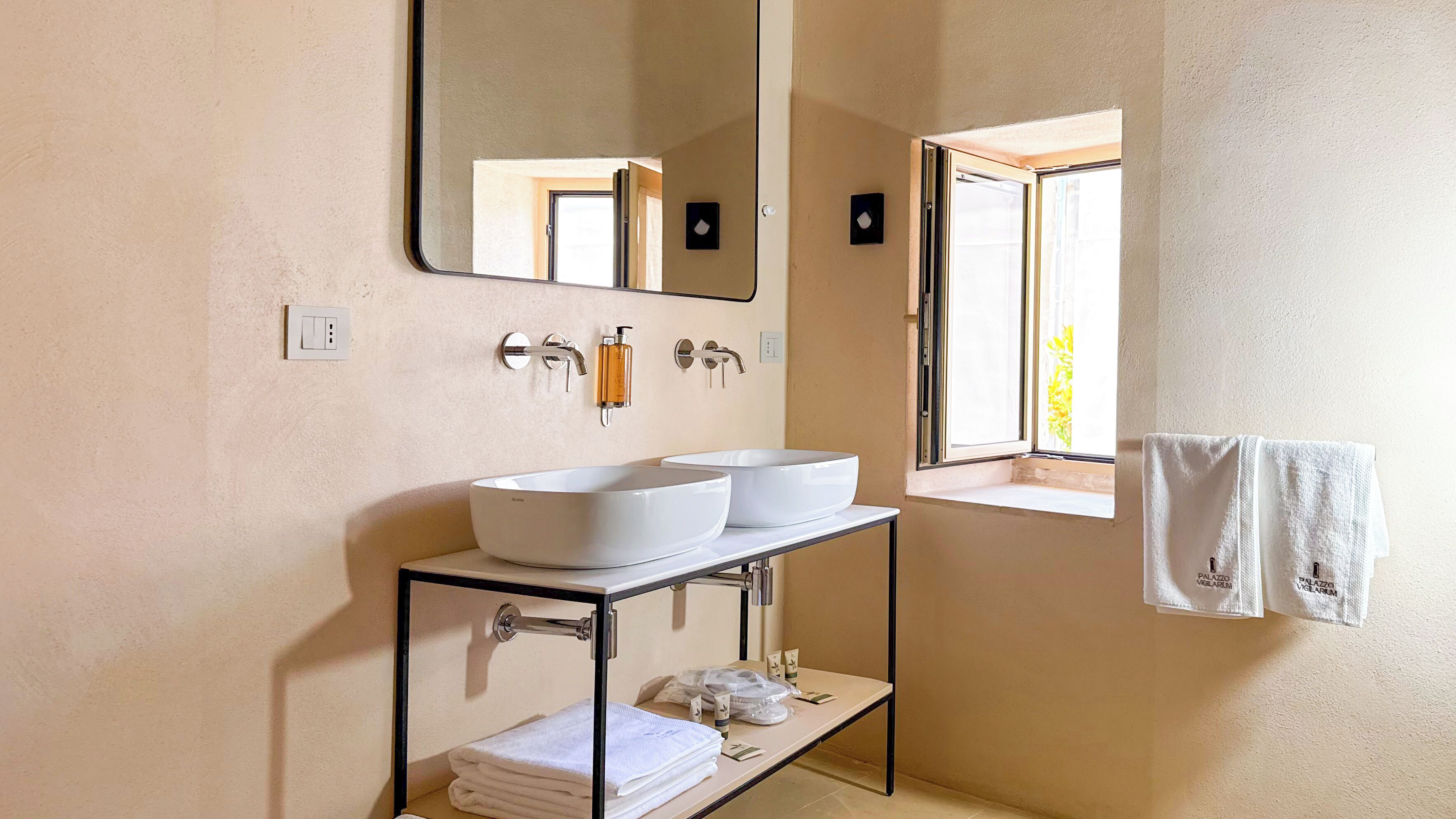 Ein Badezimmer mit zwei weißen Waschbecken, einem Spiegel, einem Fenster und einem Handtuchhalter.
