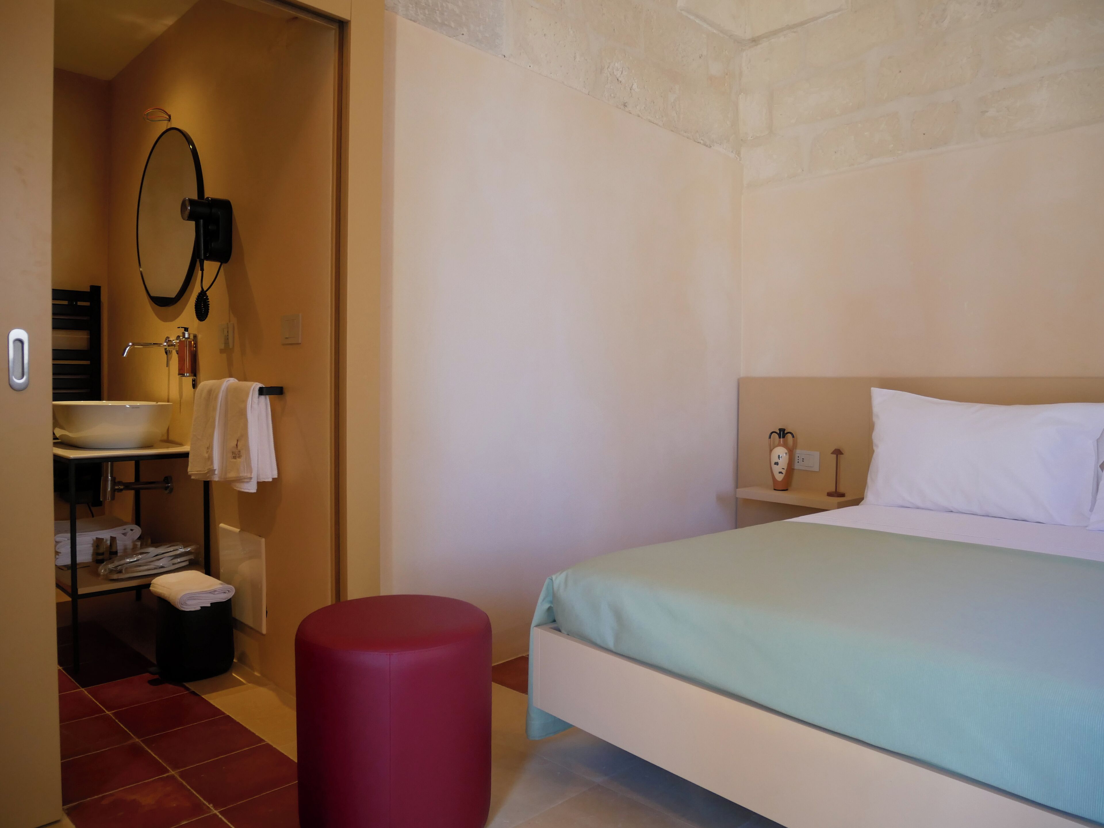 Quarto de hotel com uma cama, banheiro com pia, espelho e um banquinho vermelho.