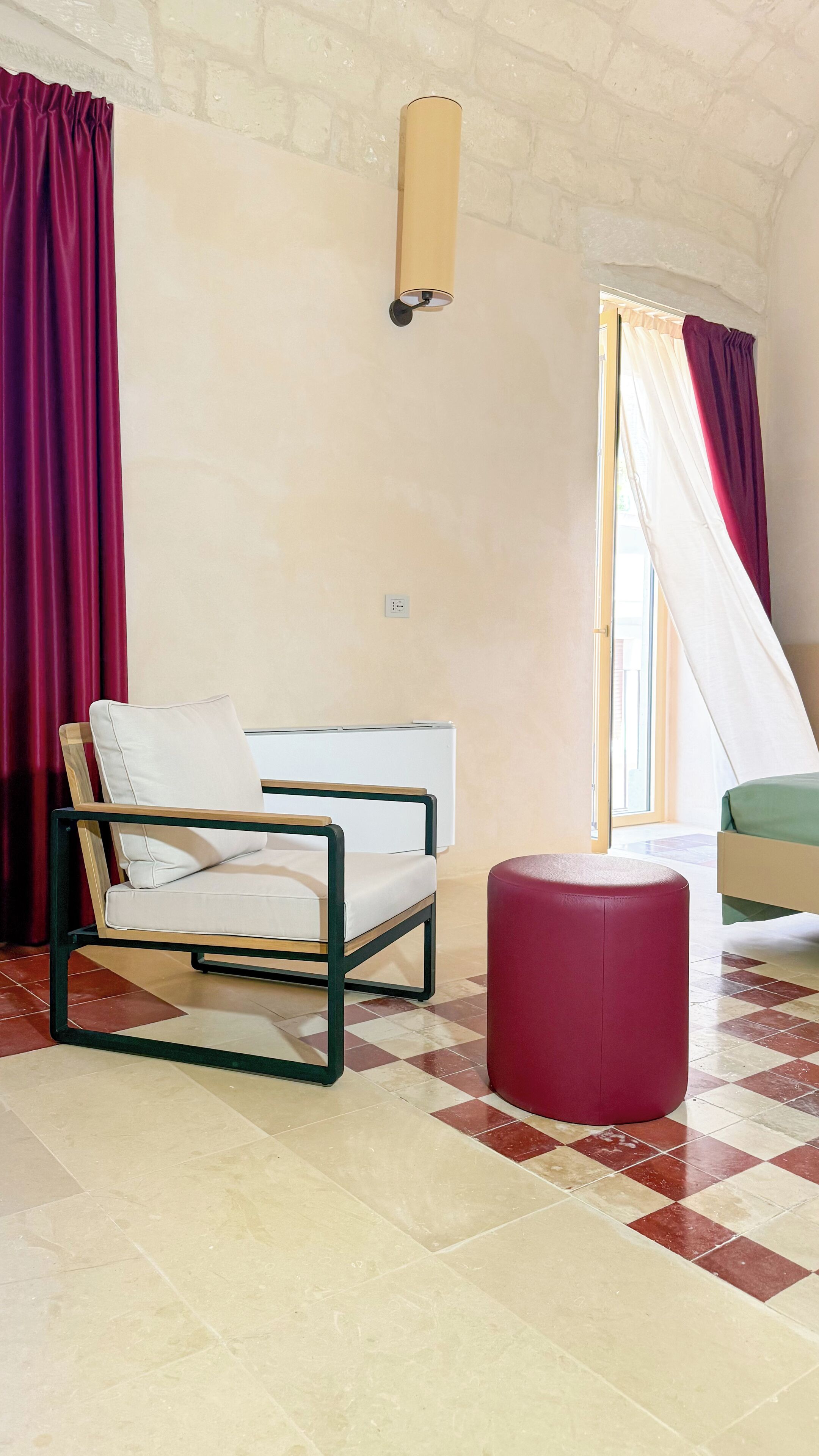 A room with a bed, a chair, a red ottoman, a wall-mounted lamp, and a window with curtains.