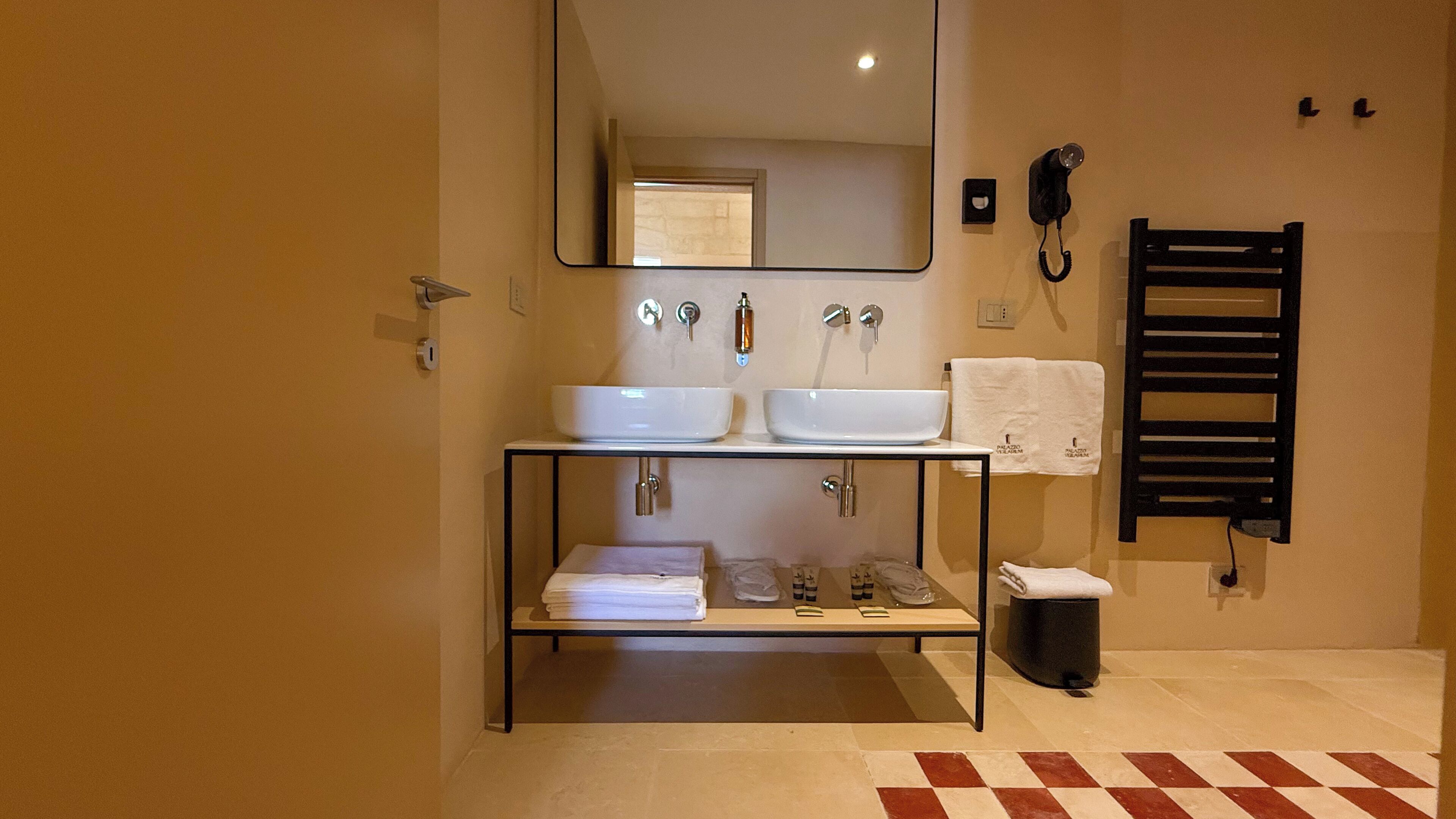 A bathroom with two sinks, a mirror, a towel rack, and a trash can.