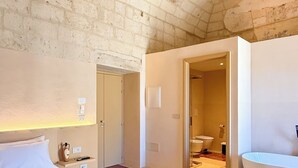 A bedroom with a bed, a bedside table, a large window, and a bathroom with a bathtub. - PALAZZO VIGILARIUM - Boutique Rooms (Uggiano la Chiesa)