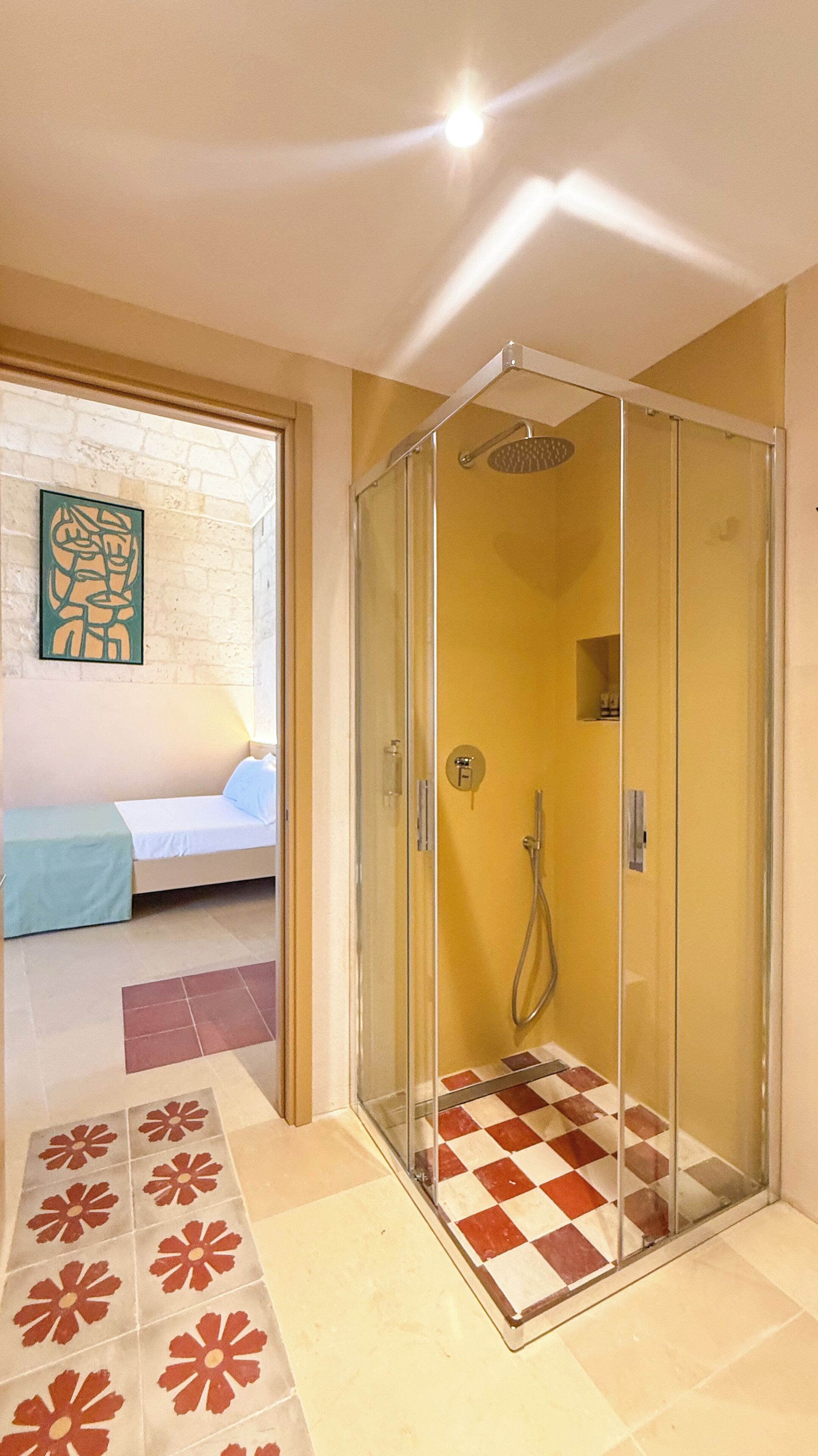 A glass-enclosed shower with a checkered floor pattern, a bed with a blue cover, and a wall-mounted artwork.