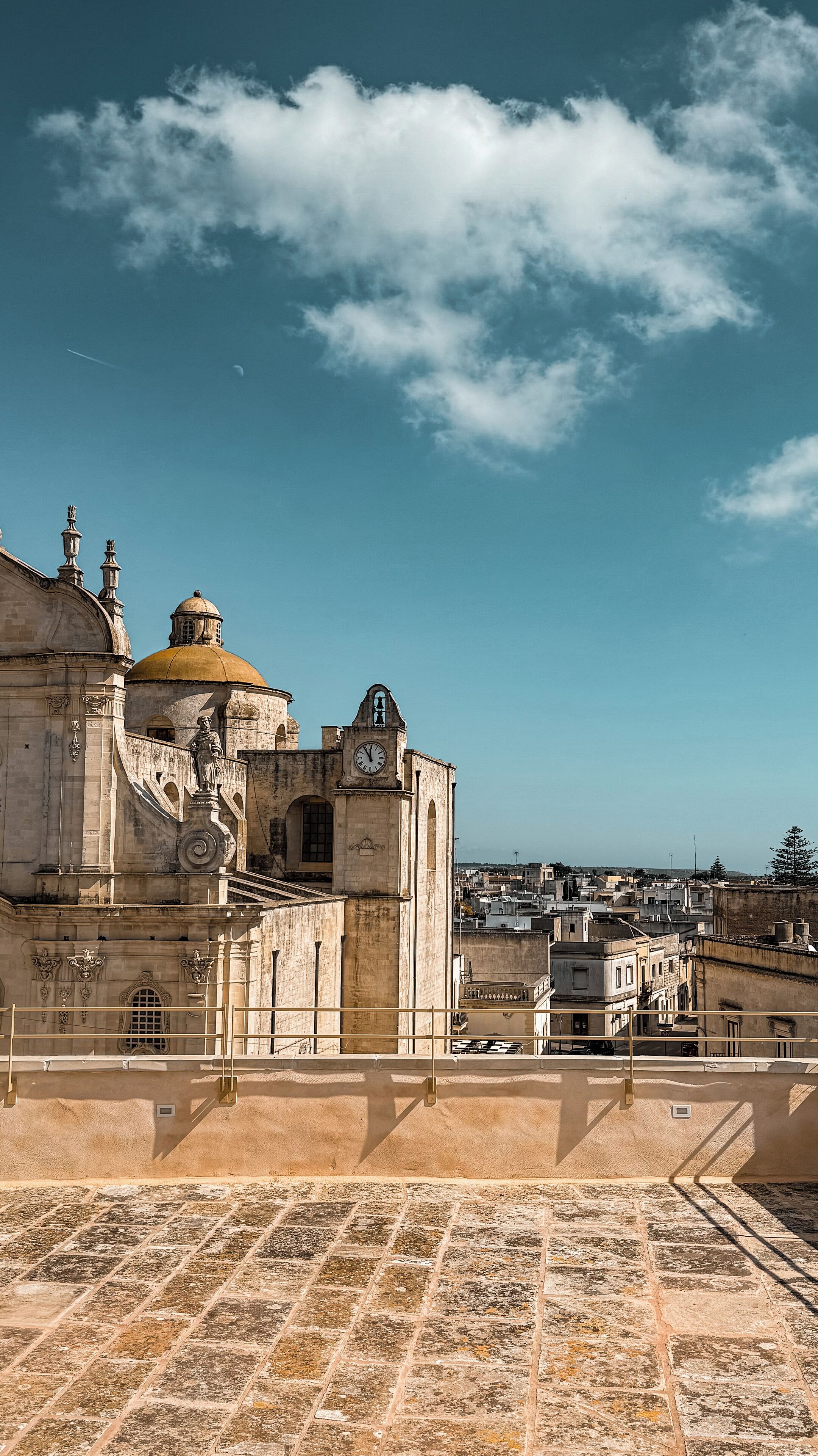 Un edificio histórico con una cúpula destacada y una torre del reloj, visto desde una terraza con suelo de piedra.