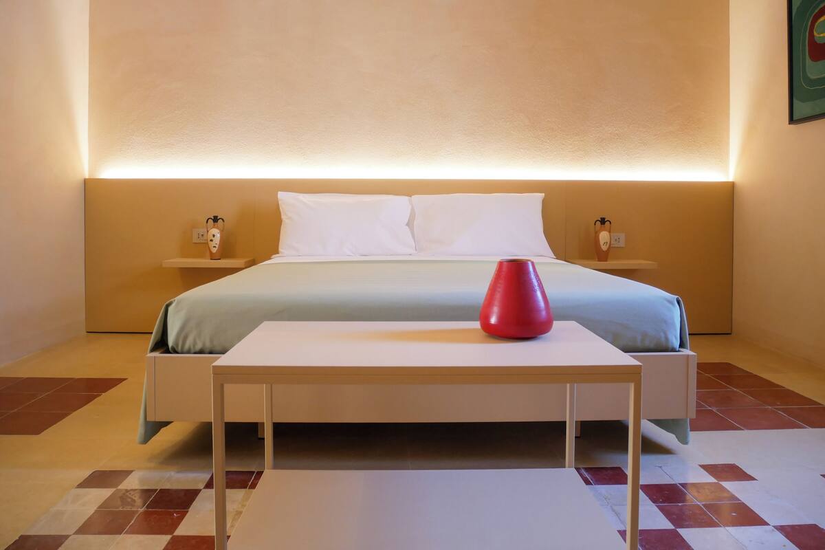Une chambre d’hôtel moderne avec un lit bien fait, une table de chevet et un vase rouge posé sur la table.