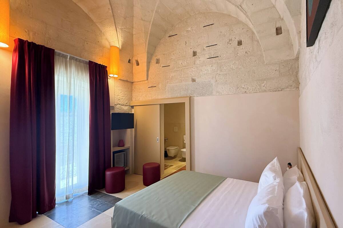 Une chambre d’hôtel avec un lit, une petite table, un tabouret rouge, une salle de bain visible à travers une porte ouverte et un mur en pierre.