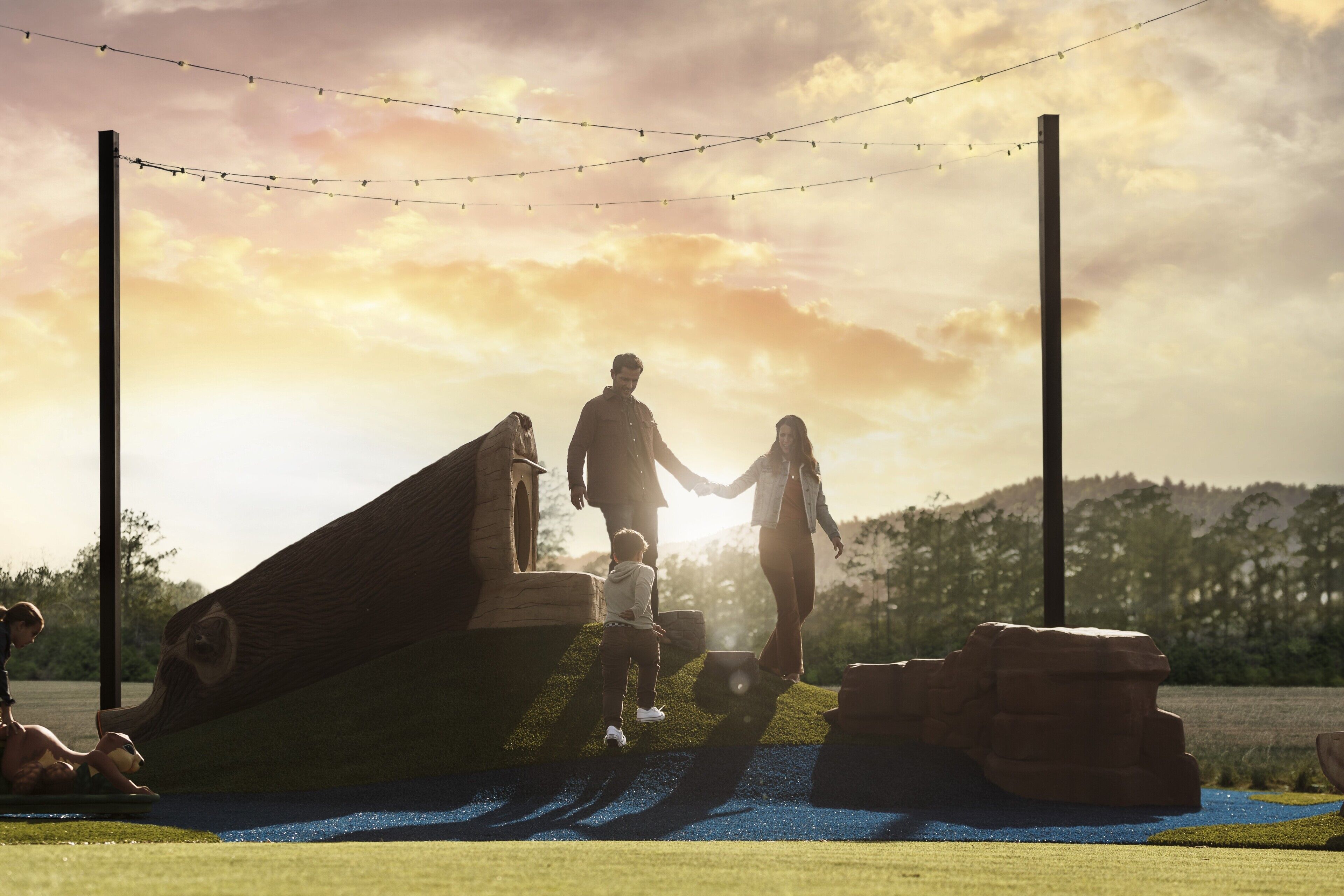 A couple holding hands on a grassy area with a wooden structure and string lights.
