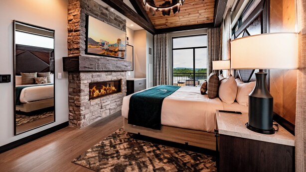 A bedroom with a stone fireplace, a bed with a view, and a large window. - Choctaw Landing Casino & Resort, Trademark Collection by Wyndham (Hochatown)