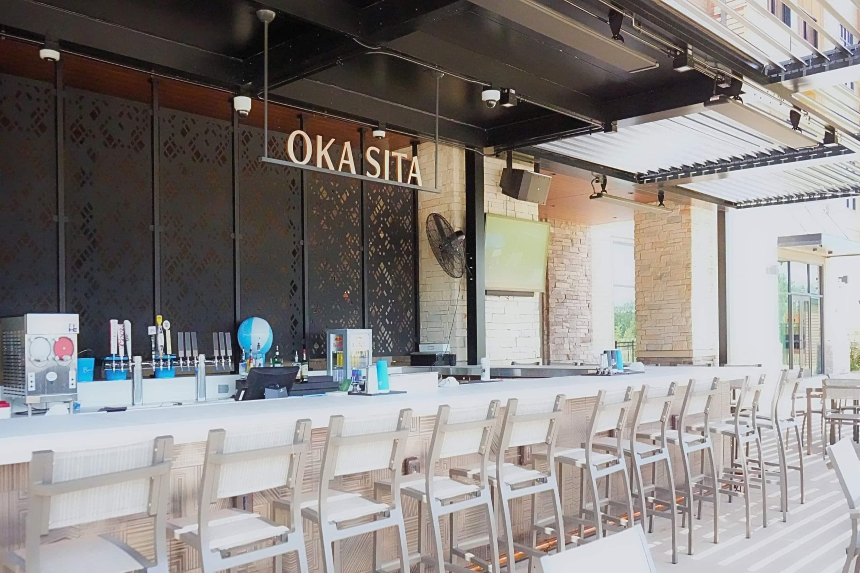 Zona de bar con un letrero que dice 'OKASITA', taburetes y una barra con diversos artículos.