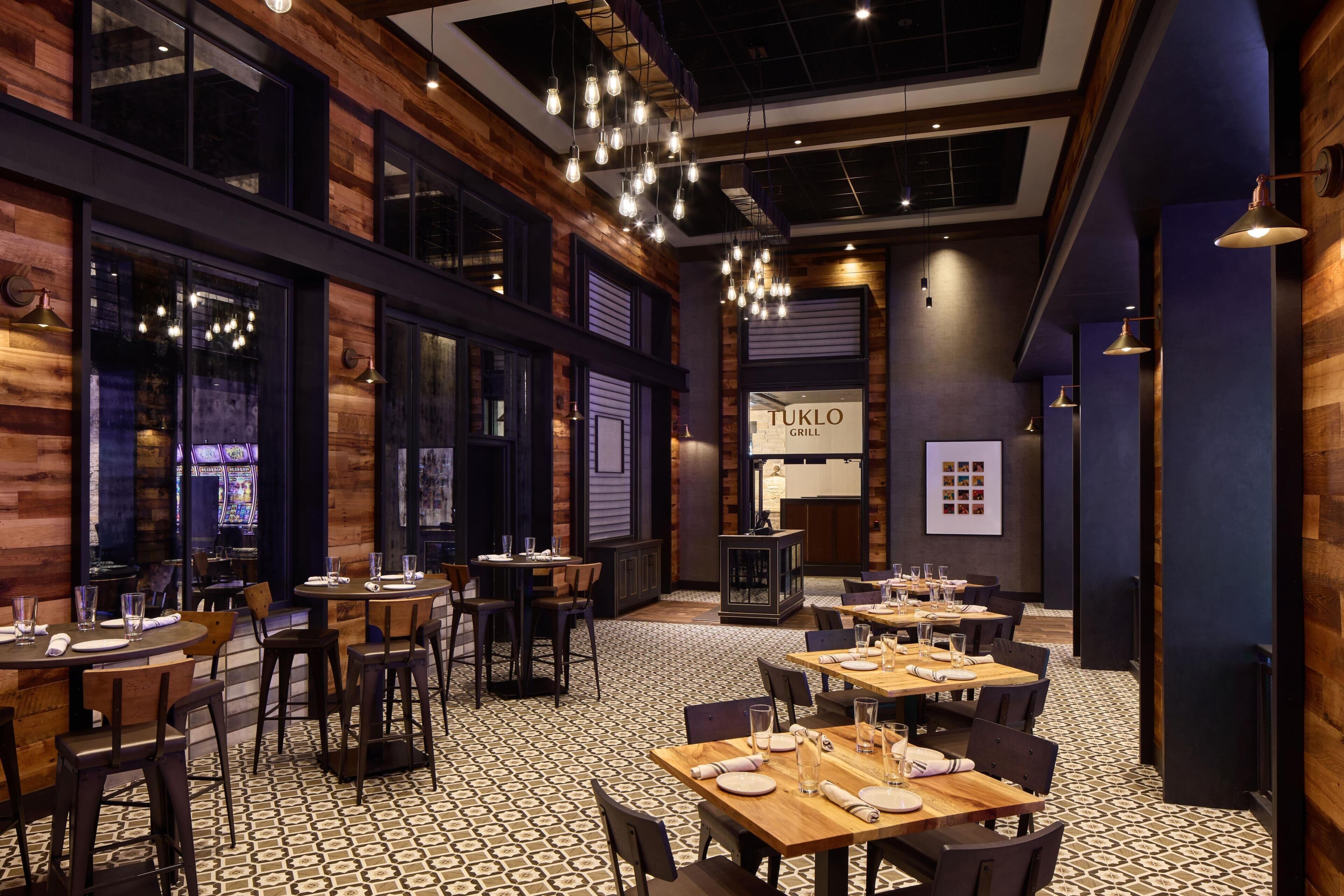 A restaurant interior with wooden walls, hanging light fixtures, and a patterned floor. Tables are set with plates and glasses.