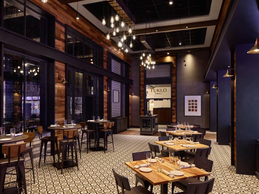 Interior de restaurante con paredes de madera, lámparas colgantes y suelo estampado. Las mesas están preparadas con platos y vasos.