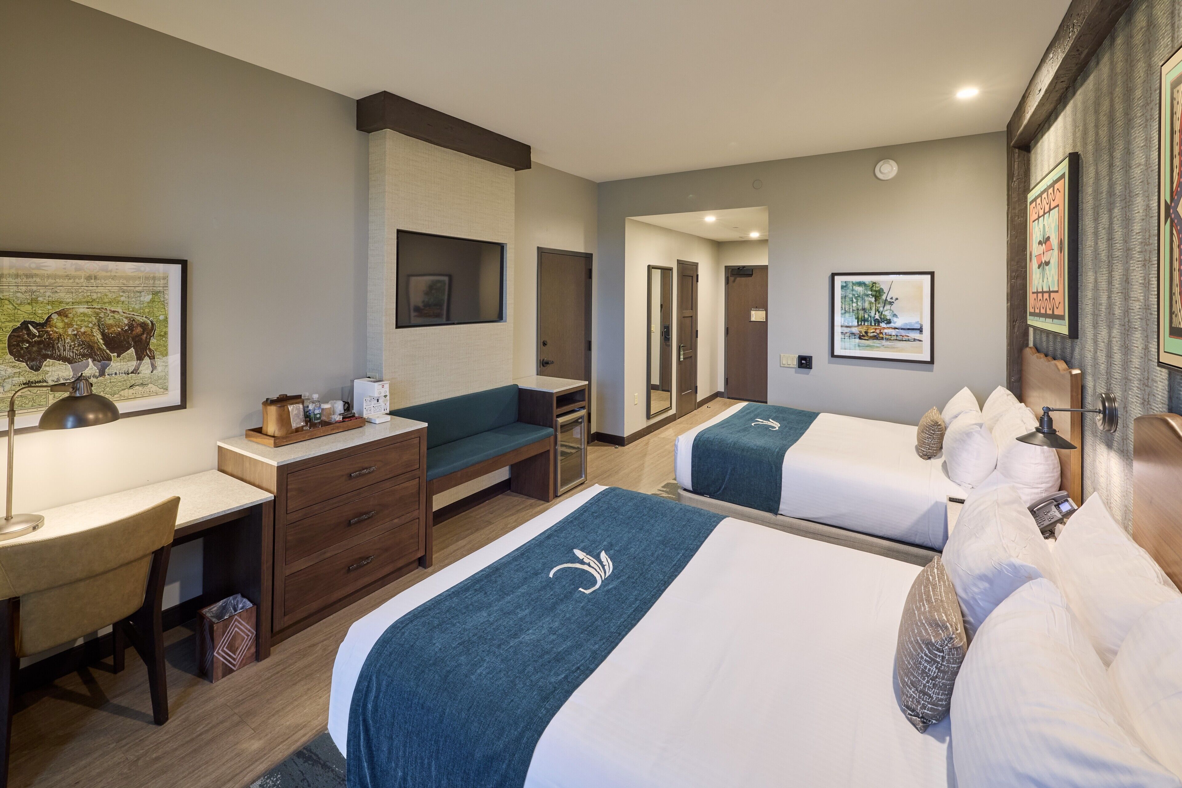 Habitación de hotel con dos camas, un escritorio, una silla, un aparador y obras de arte en las paredes.