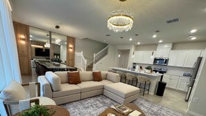 A modern living room with a sectional sofa, a dining table with chairs, a round coffee table, and a kitchen area with white cabinets and stainless steel appliances. - Family Friendly 5Bd Near Disney Windsor Cay 16136 (Clermont)